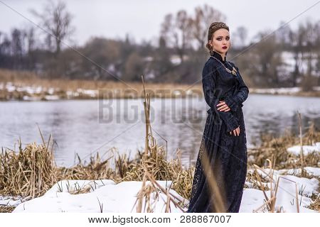 Young Caucasian Brunette Female In Art Dress With Necklace And Crown Posing Against River Outdoors. 