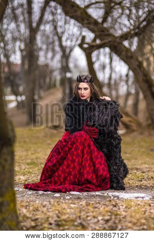 Beautiful Princess In Red Dress And Black Fur With Crown And Her Dog In Forest During Early Spring. 