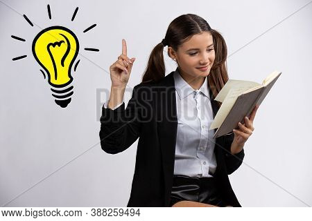 Beautiful Teenage Girl Reading Book And Pointing Up With Finger On Light Bulb, Isolated On White