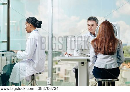 Serious Experienced Ophthalmologist Checking Eyesight Of Young Woman, His Female Colleague Workng On