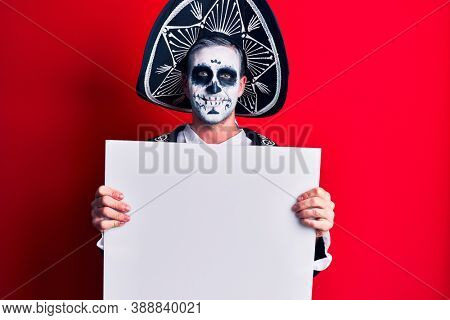 Young man wearing mexican day of the dead costume holding blank empty banner looking positive and happy standing and smiling with a confident smile showing teeth 