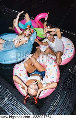 Angle View Of Woman In Party Headband With Blow Tickler Swimming On Ring Near Friends In Pool At Nig