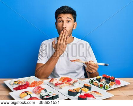 Young handsome man eating sushi sitting on the table covering mouth with hand, shocked and afraid for mistake. surprised expression 