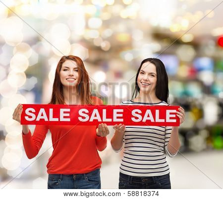 shopping, sale, mall and gift sconcept - two smiling teenage girls with sale sign at shopping mall