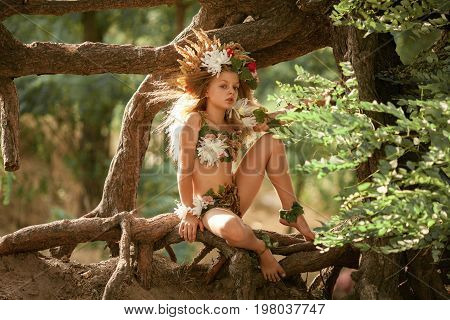 Beautiful little girl in image of nymph dryad with floral head wreath sits on tree roots in magical fairytale forest .