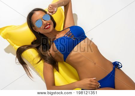 Sexy girl wearing sunglasses and blue bicini lying on yellow swimming air mattress isolated