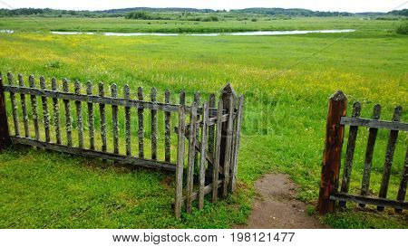 An open gate in a wooden fence and a green meadow beyond it, the path in the frame . Cloudy summer or late spring outside the city