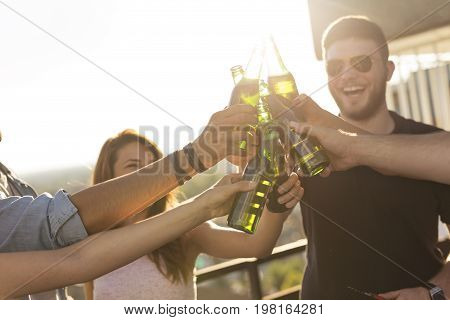 Group of young friends having fun at rooftop party drinking beer making a toast and enjoying hot summer days. Selective focus on the beer bottles and hands