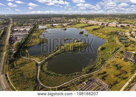 Aerial view of a lake and peninsula near a community center and townhomes with walking paths and bridge in The Glen, Glenview, Illinois.