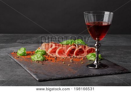 A wineglass filled red wine, salted and raw balyk, spicy basil on a black background. Prosciutto and semi sweet wine. A glass of wine and a pile of cut ham on a black background. Copy space.