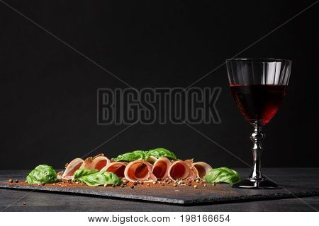 A wineglass filled red wine, salted and raw balyk, spicy basil on a black background. Prosciutto and semi sweet wine. A glass of wine and a pile of cut ham on a black background. Copy space.