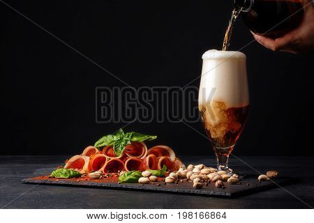 A man's hand add alcohol beer in a transparent glass and near a cured prosciutto or balyk with pistachios, a spinach and a light beer with foam on a black background. Copy space.