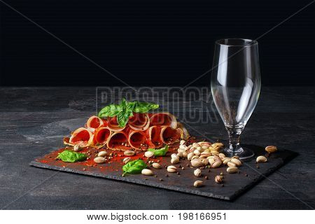 A composition of an empty glass for beer, pistachios and a plate of perfectly sliced balyk with a spinach on a black background. Prosciutto with a basil and a little bit beer in a glass. Copy space.