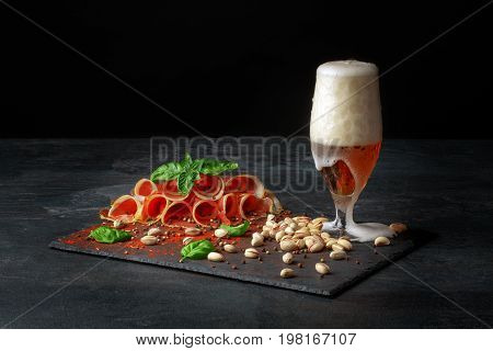 A composition of a glass of dark beer, pistachios and a plate of perfectly sliced balyk with spinach on a dark background. Cured prosciutto with a basil and a goblet of alcoholic beer with foam.