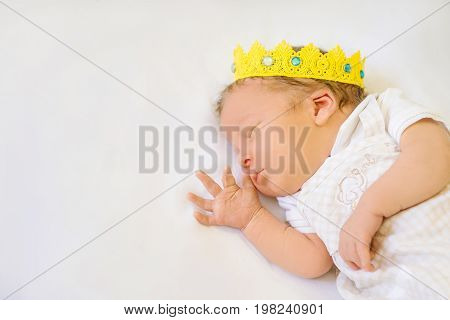Portrait of a 12 day old newborn baby boy wearing a gold crown. He is sleeping on a beige flokati rug with his hands behind his head.