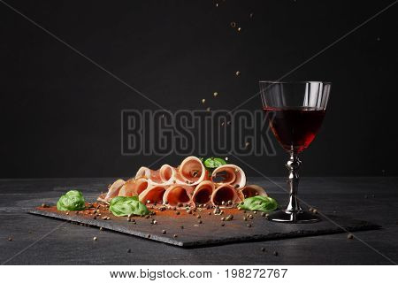 A glass of red wine and thinly sliced balyk on the saturated black background. Dry red wine, traditional Italian snacks, basil and peppercorns on the table. Copy space.