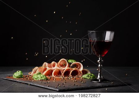 A glass of red wine and thinly sliced balyk on the saturated black background. Dry red wine, traditional Italian snacks, basil and peppercorns on the table. Copy space.