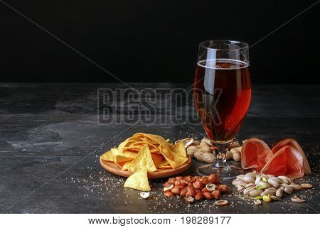 A beautiful composition of different snacks and a pint of saturated brown beer on a black table background. Peanuts, pistachios and a plate of chips. A beer with a perfectly sliced balyk. Copy space.