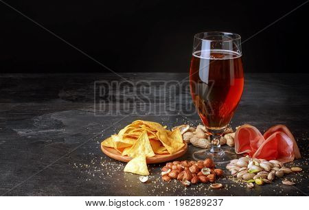 A beautiful composition of different snacks and a pint of saturated brown beer on a black table background. Peanuts, pistachios and a plate of chips. A beer with a perfectly sliced balyk. Copy space.