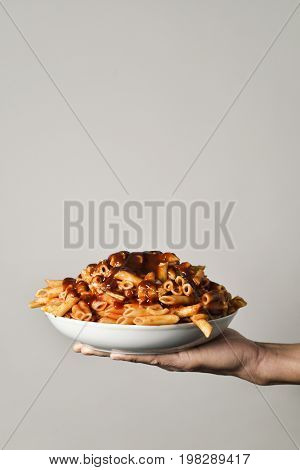 closeup of a young caucasian man holding a plate with an assortment of different cooked pasta served with tomato sauce in his hand, against an off-white background with a blank space on top