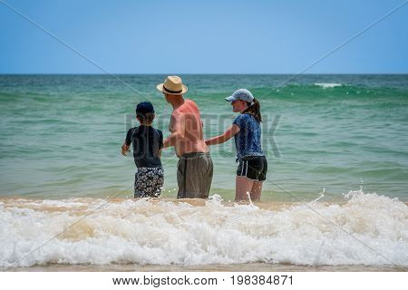 Three people bathing in waves.
Albufeira Portugal - July 17 2017: Seen from behind a caucasian male in sunhat with a young boy and girl standing in water with waves and looking at the horizon. Bathing at a public beach at the Algarve coast.