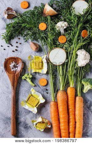Raw vegetables and bouillon cubes for cooking soup. Young carrot with haulm, broccoli, cauliflower, onion, garlic, salt, pepper, spoon over gray concrete background. Top view. Dinner cooking concept