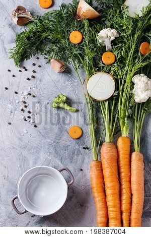Raw vegetables for cooking soup. Ingredients young carrot with haulm, broccoli, cauliflower, onion, garlic, salt pepper, empty pot over gray concrete background. Top view. Dinner cooking concept