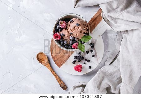 Homemade chocolate ice cream with frozen berries blueberry, raspberry, mint served in white bowl with olive wood spoon, marble serving board, textile over gray concrete background. Top view with space