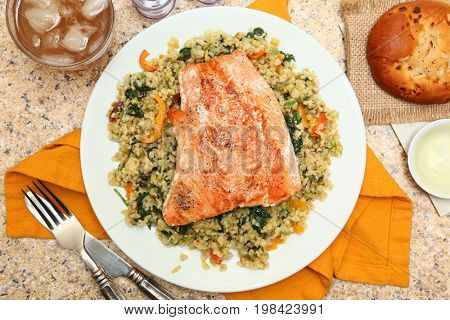 Salmon with Cracked Freekeh Salad at table with spicy salsa verde, iced tea, lemon and an onion roll.