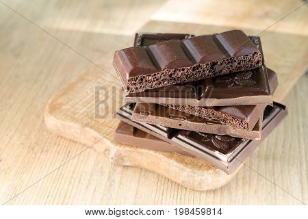 Different broken chocolate bars stacked on a wooden board. Chocolate lit by the sun. Place for text.