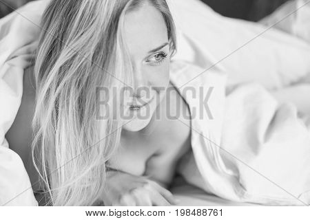 Black & white portrait of Caucasian ladyposing with happy smile in morning bed.Young attractive blue eyed female with long blond hair biting liplooking playfully sideways.Health and beauty concept.