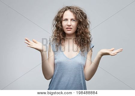 Unsure confused doubtful European woman with long curly hair in grey casual t-shirt shrugging shoulders in questioning gesture of uncertainty and keeping mouth openedhaving confused clueless look.