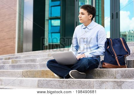 Portrait of thoughtful teenage boy sitting on school stairs outdoors using laptop