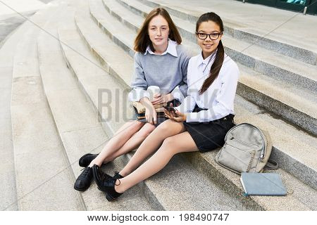 Portrait of two pretty schoolgirls looking at camera while sitting on stone stairs outdoors