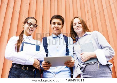Portrait of three smiling schoolchildren holding books looking at camera standing against bright orange wall