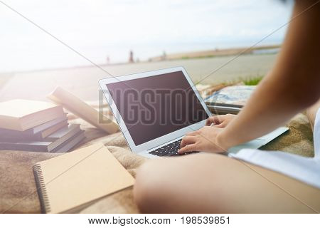 Outdoor summer shot of college student girl keyboarding on laptop pc working on course paper sitting alone on the riverbank with many books and copybook. People education and technology concept