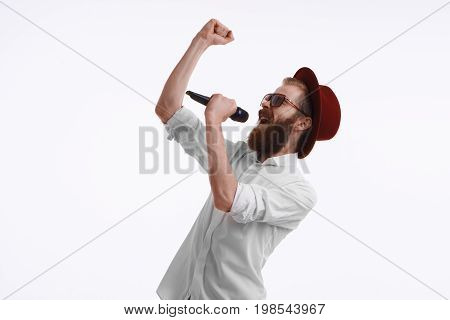 Emotional adult male entertainer actor or pop-singer wearing white shirt stylish hat and sunglasses holding wireless microphone speaking or singing in studio. Show entertainment and performance