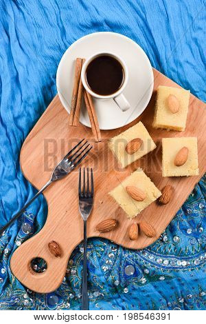 Semolina halva squares with almonds served with coffee on blue embroidered cloth flat lay. Indian sweets overhead view