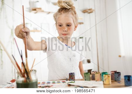 Indoor shot of European cute creative child with hair bun snd blue eyes occupied with drawing. Little blonde girl wearing white t-shirt busy with painting, deeping brush into water, drawing new masterpiece for her parents.
