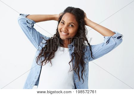 Studio shot of good-looking attractive young Afro-American woman in denim shirt and white t-shirt playing with her long wavy hair, smiling broadly with her perfect white teeth to the camera. Dark-skinned cheerful female posing on white background.