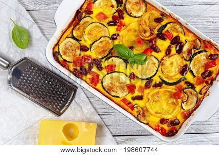 delicious zucchini casserole with red bell pepper red bean ham and cheese in gratin dish on old wooden table with piece of cheese grater on papel view from above close-up