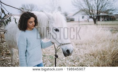 Attractive woman in soft blue jamper and jeans walking with a amazing white horse outdoors on the field in a bright sunny day. Close up