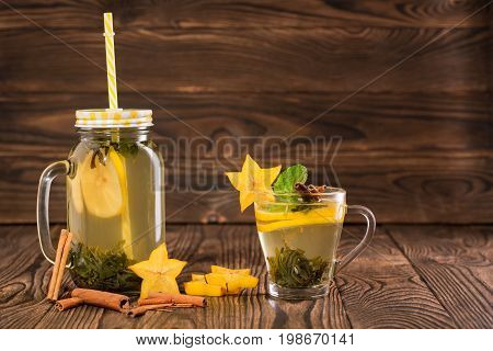 Composition of mason jar of lemon tea with yellow straw and a cup of green tea with mint and tropical carambola on a light wooden background. Healthy homemade tea with cinnamon and fruits. Copy space.