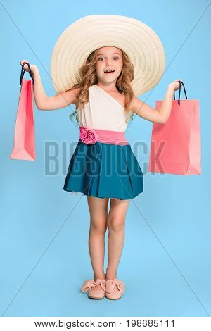 The cute little caucasian brunette girl in hat holding shopping bags on blue studio background