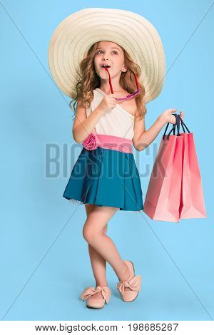 The cute little caucasian brunette girl in hat holding shopping bags on blue studio background