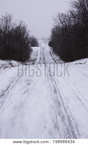 Vertical of a long snow covered country road framed by bare trees and leading to a distant tree line in Akwesasne, New York on an overcast blustery day in February.