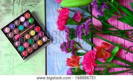 Colorful makeup palette and beautiful flowers on multicolored wooden background