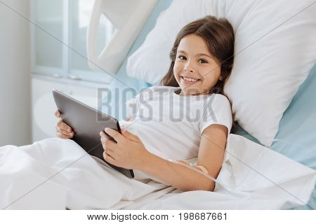 Modern igeneration. Cheerful elated cute girl lying on the hospital bed and smiling while holding a tablet