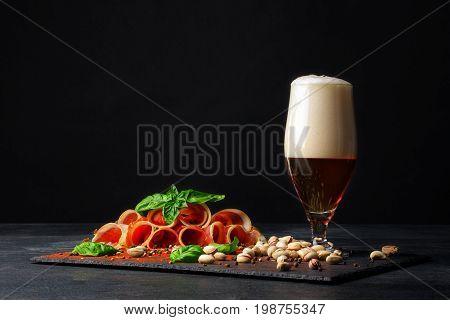 A beautiful composition of different snacks and a pint of saturated brown beer with thick froth on a black background. Pistachios and a plate of perfectly sliced balyk pieces. Copy space.