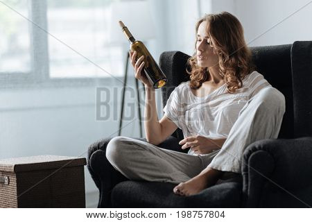 Bottle of wine. Sad unhappy cheerless woman holding a bottle of wine and looking at it while sitting in the armchair